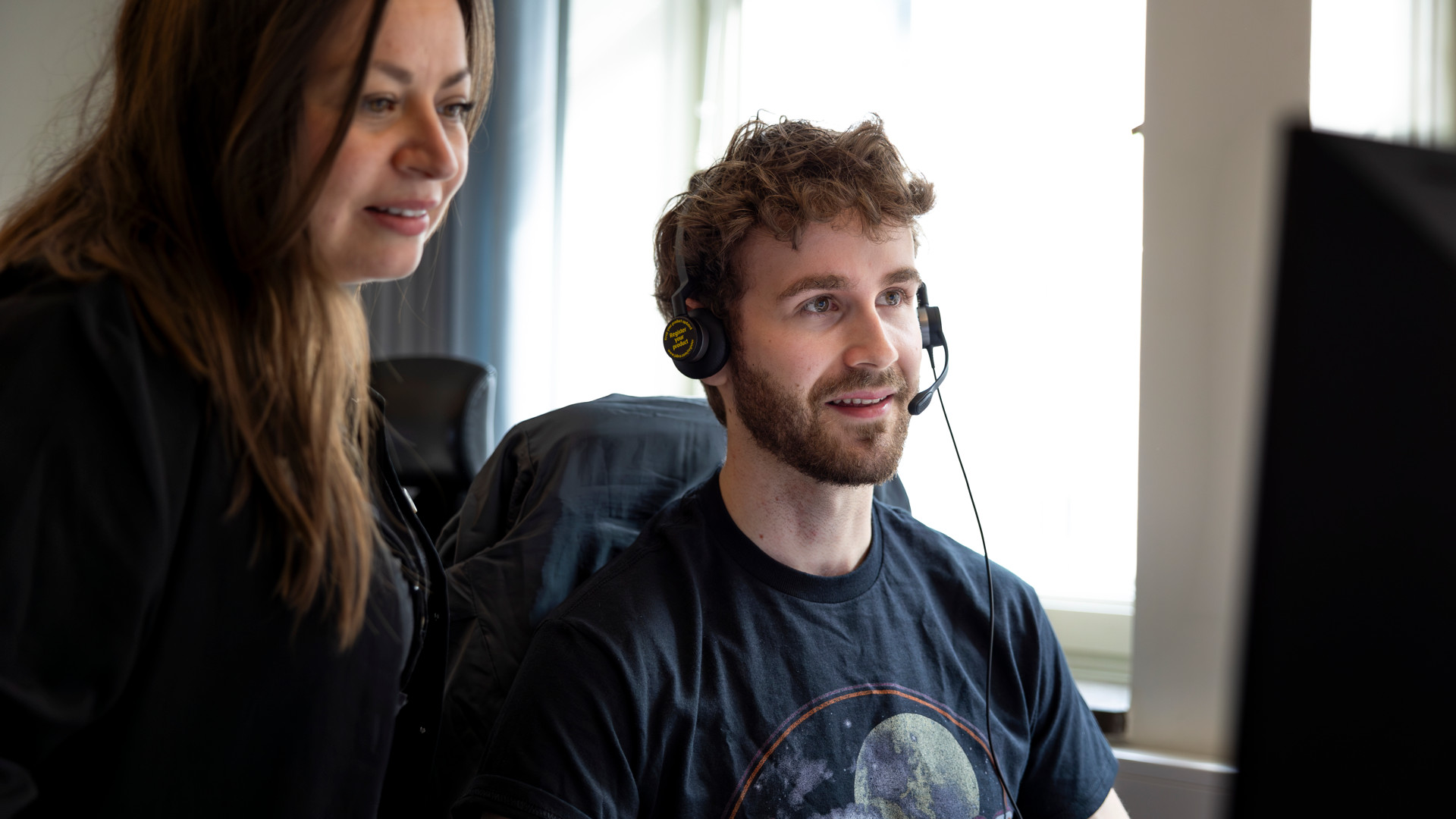 En man med headset och skägg sitter vid en dator och tittar på skärmen med ett leende. En kvinna står bredvid honom och tittar också på skärmen, båda ser engagerade ut.