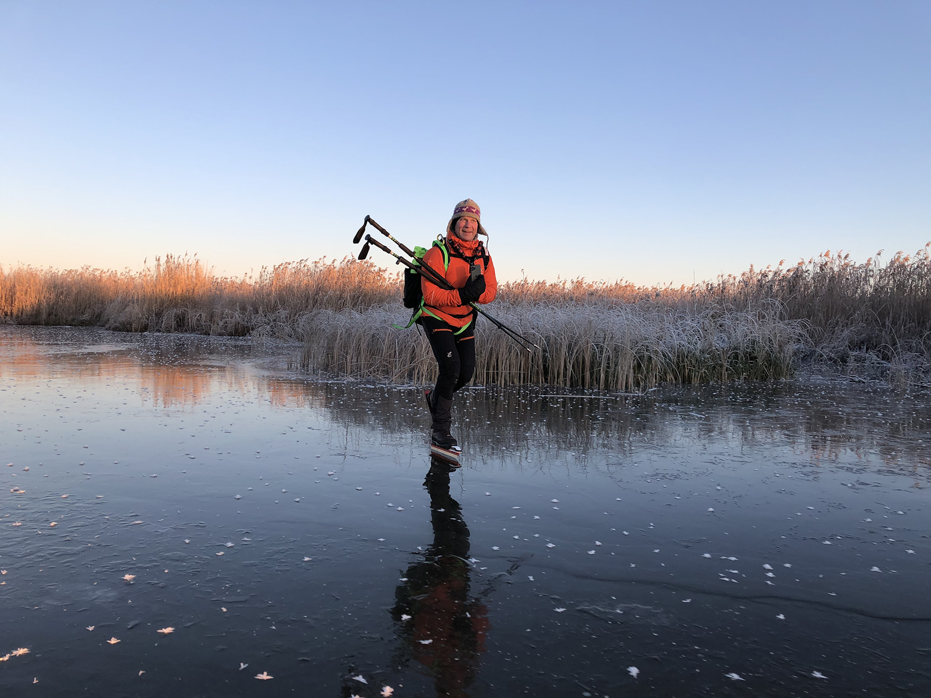 En person klädd i orange jacka och ryggsäck åker långfärdsskridskor på en frusen sjö vid soluppgången. Vassen i bakgrunden är frosttäckt och himlen är klar.