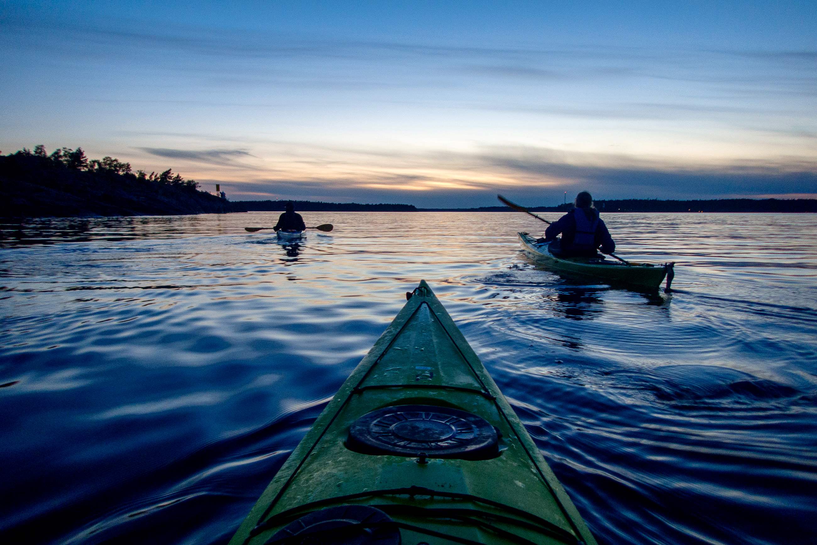 Två personer paddlar kajak på en lugn sjö vid solnedgången, med silhuetter av skog och en färgglad himmel i bakgrunden.