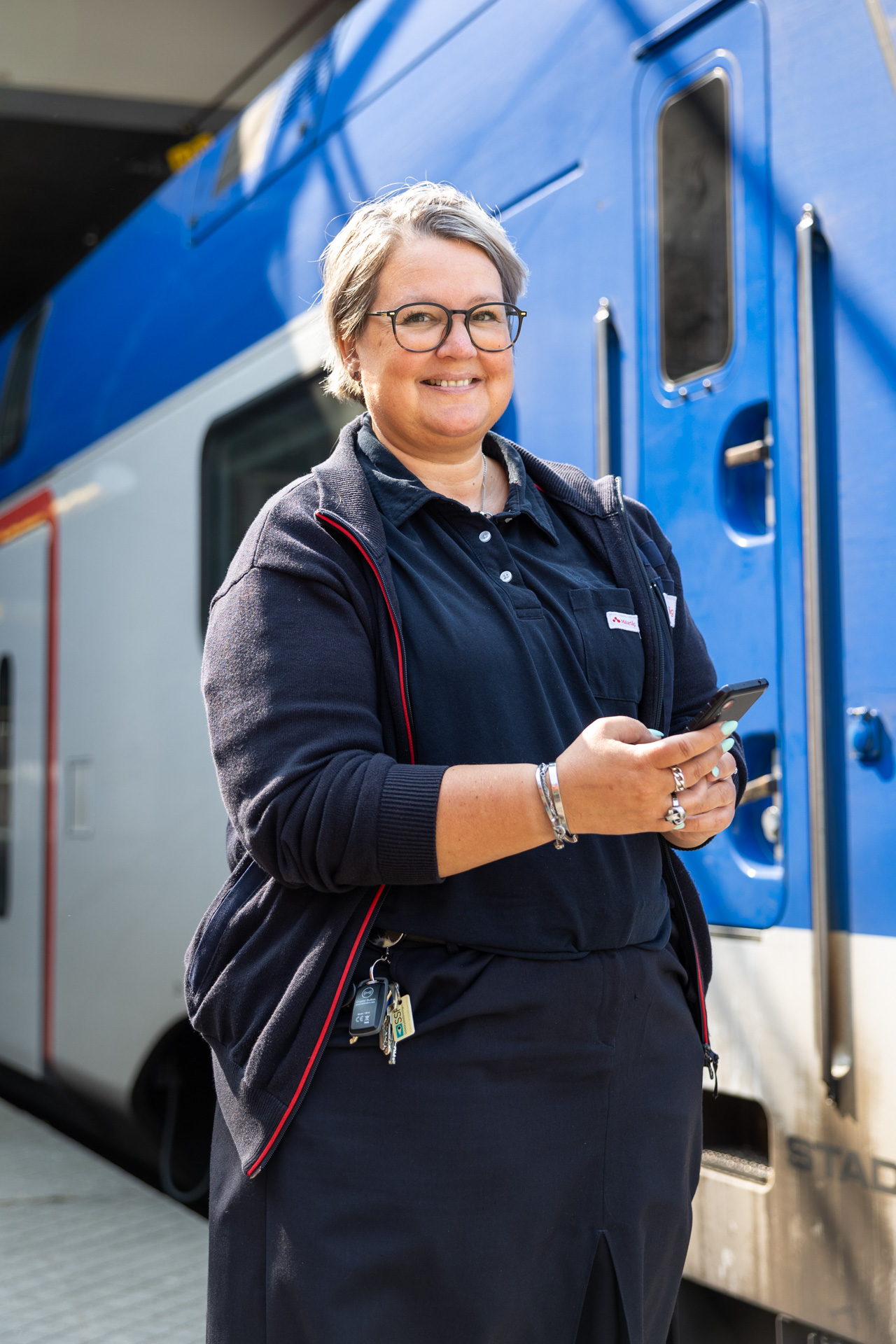En kvinnlig tågvärd med glasögon och kort grått hår står framför ett blått och vitt tåg, leende och håller en mobiltelefon i handen.