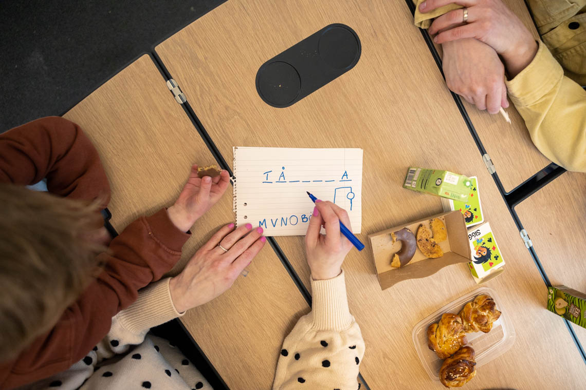 En grupp människor spelar hänga gubbe vid ett bord. På bordet ligger ett papper med spelet, pennor, kakor, bullar och dryck i små förpackningar.