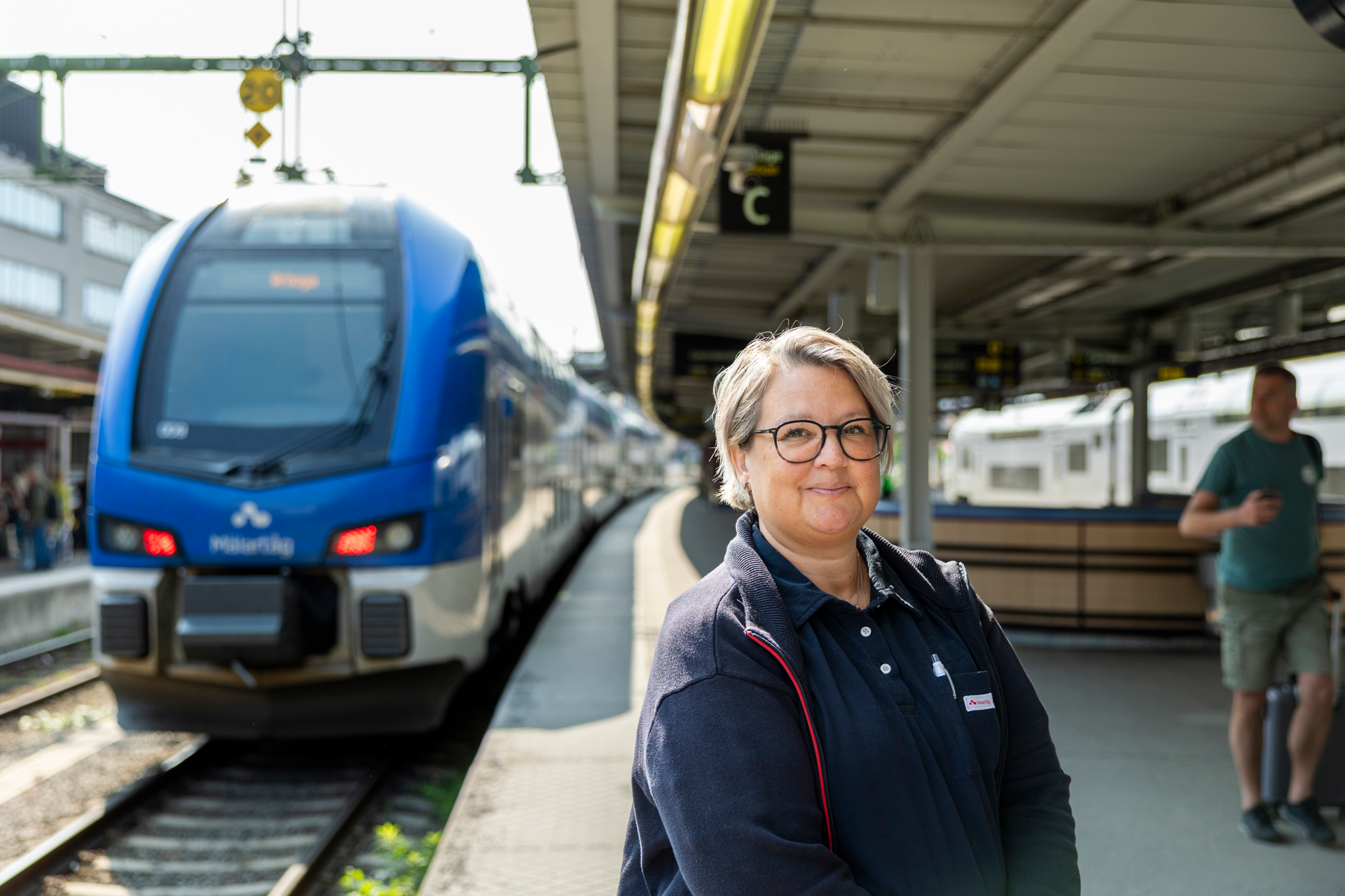 En kvinnlig tågvärd står framför ett tåg på en tågstation