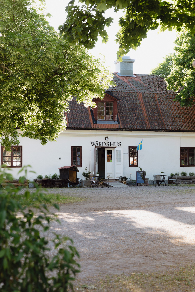 Ett charmigt gammalt värdshus med vit fasad och tegeltak, omgivet av grönskande träd. Utanför syns några utemöbler och en svensk flagga.