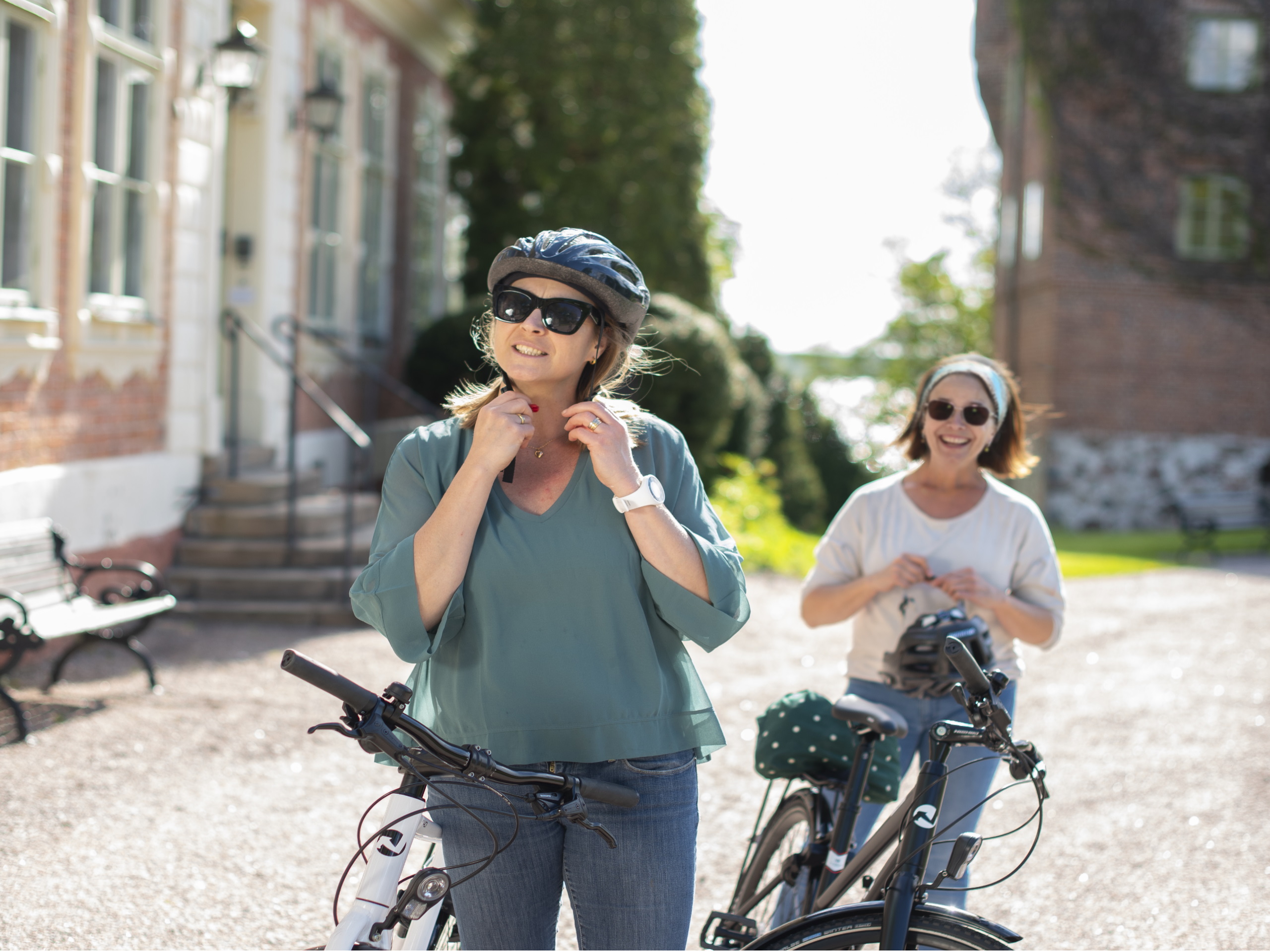 Två kvinnor spänner på sig cykelhjälm vid sina cyklar utanför ett slott. 