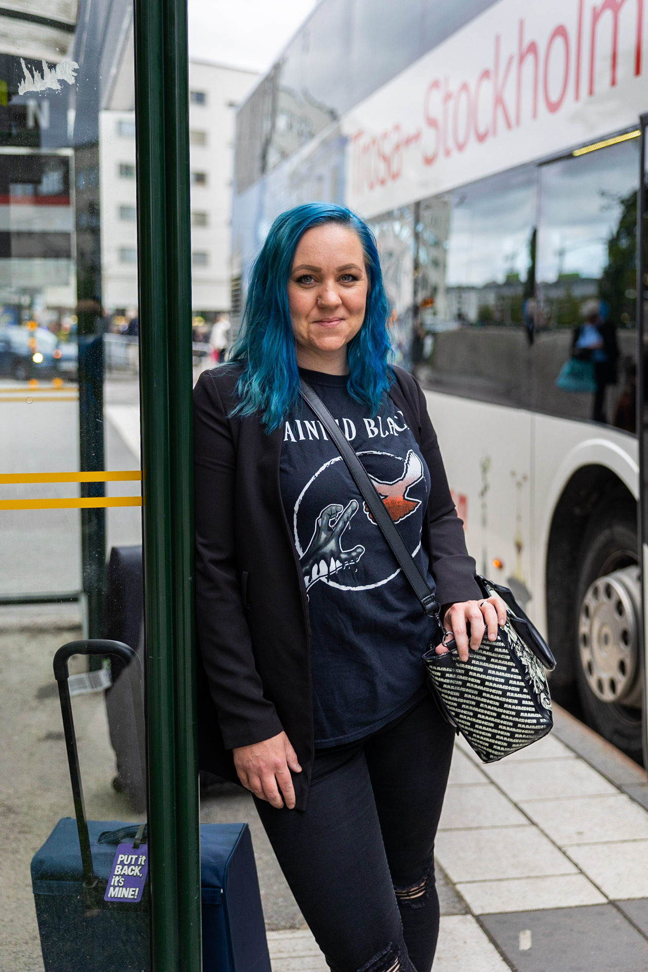 En kvinna med blått hår står vid en busshållplats bredvid en buss märkt "Trosa-Stockholm". Hon håller en liten väska och har en resväska vid sidan.