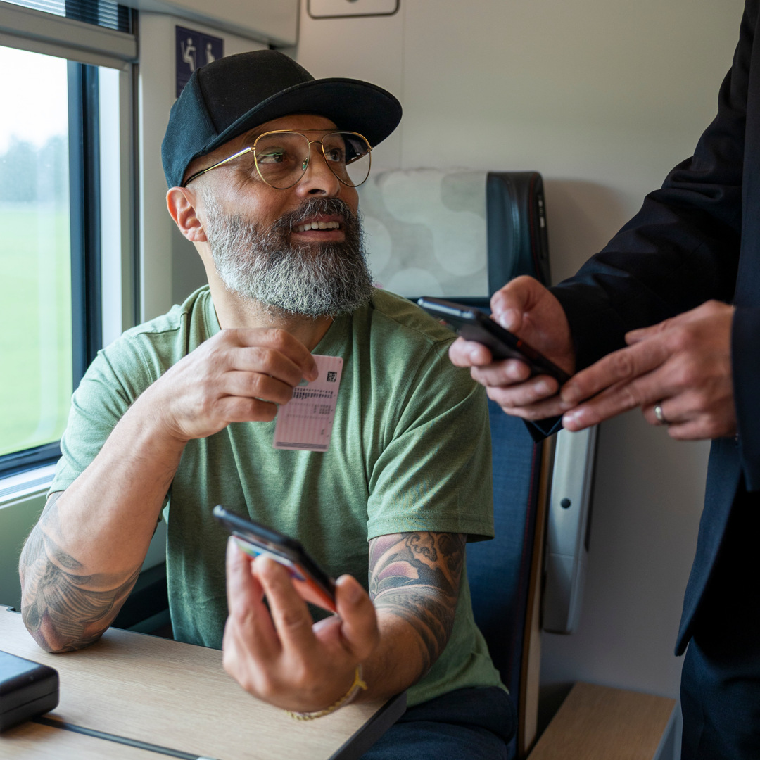 En man sitter vid ett bord med en mobiltelefon och blir viserad av tågvärd. Håller upp sin legitimation.